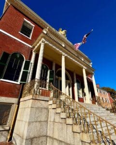 Angled view of the Salem Custom House in Salem, Massachusetts, featuring its golden eagle, grand staircase with ornate railings, and red brick facade under a bright blue sky.