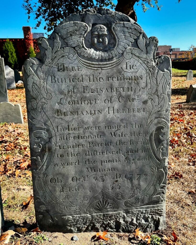 Gravestone of Elisabeth Herbert in Salem Massachusetts's Old Burying Point cemetery, featuring a carved angel at the top, intricate floral details, and an inscription honoring her as a devoted wife and mother, who passed in 1772 at age 55.