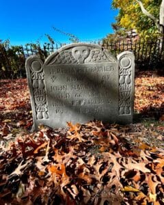 Close-up of John Marston Senior's 1681 gravestone in Salem Massachusetts's Old Burying Point cemetery, featuring a winged skull carving symbolizing mortality and surrounded by vibrant autumn leaves under a bright blue sky with trees and an iron fence in the background.