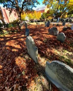 Weathered gravestones surrounded by a bed of red and orange fallen leaves at Old Burying Point Cemetery in Salem Massachusetts, with sunlight filtering through trees and colorful autumn foliage.