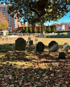 Gravestones at the Old Burying Point cemetery in Salem Massachusetts, shaded by a large tree with branches extending over the historic burial ground, surrounded by colorful autumn leaves on the ground.