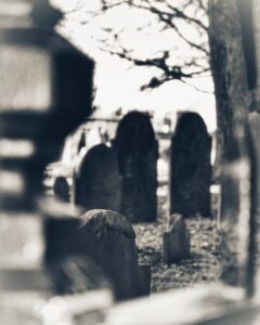 A black-and-white photo showing gravestones at Old Burying Point Cemetery in Salem, Massachusetts, framed through a metal fence. The headstones stand quietly beneath bare tree branches.