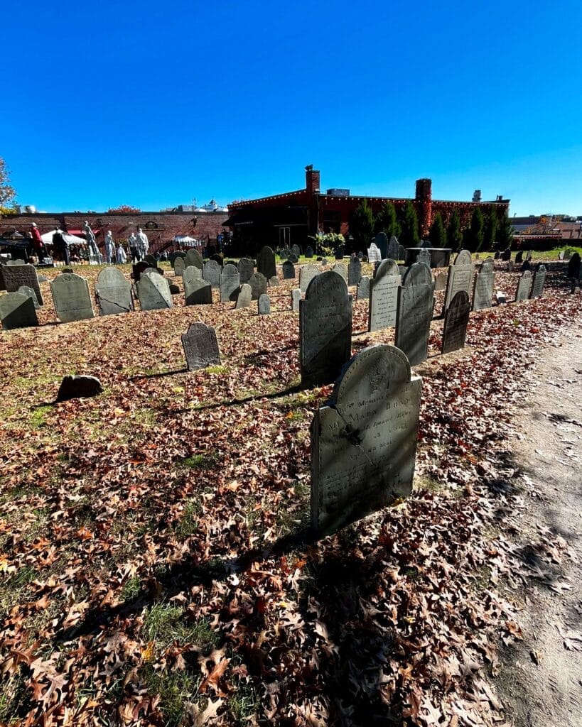 Gravestones in the Old Burying Point cemetery in Salem Massachusetts, surrounded by fallen autumn leaves, with a backdrop of a bright blue sky and nearby brick buildings.