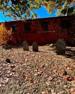 Gravestones at the Old Burying Point cemetery in Salem Massachusetts, set against a red brick wall covered with vibrant, autumn-colored vines under the canopy of a large tree.