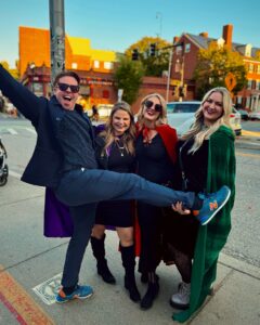 A cheerful group of four friends standing on the sidewalk in Salem, Massachusetts. One person lifts their leg in a playful pose, while the others smile brightly, wearing colorful Halloween-themed capes. The background features historic brick buildings typical of Salem's architecture.