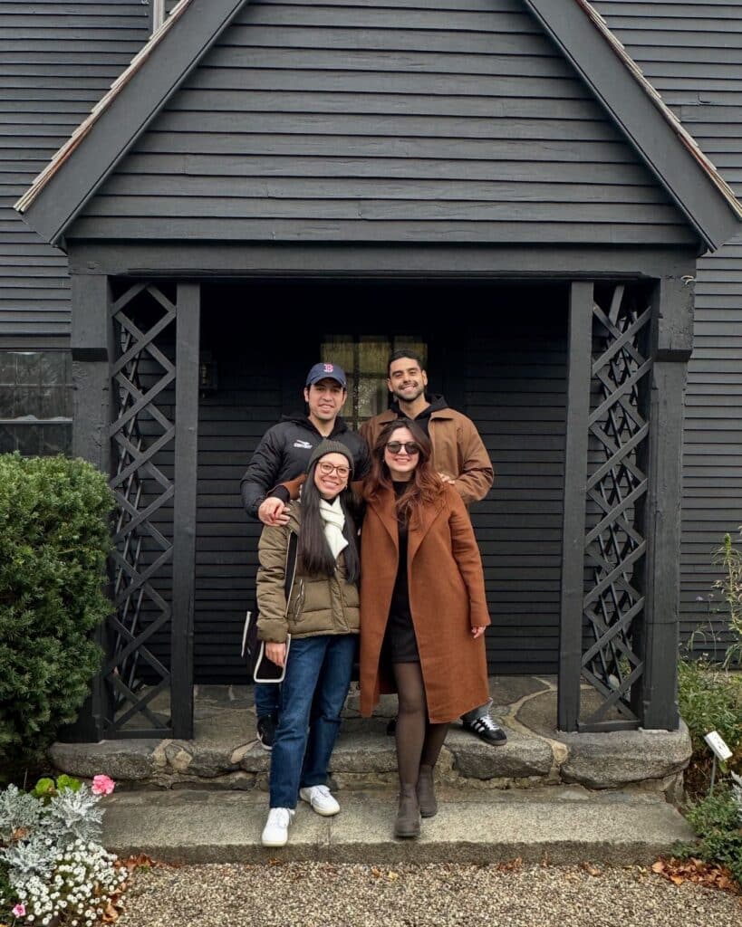A group of four people stands smiling in front of the entrance to the House of the Seven Gables in Salem, Massachusetts. The historic black wooden house with lattice details and stone steps serves as the backdrop.