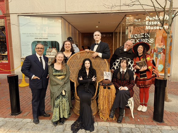A group of eight people dressed as characters from the Addams Family standing in front of the Peabody Essex Museum in Salem, Massachusetts. The group includes Morticia, Gomez, Wednesday, Uncle Fester, and other characters, posing together on the brick sidewalk.