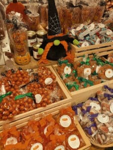 A display of various Halloween-themed candies at Ye Olde Pepper Companie in Salem, Massachusetts. The table is filled with colorful wrapped candies, pumpkin-shaped treats, and jars of sweets. A witch hat decoration with orange braids is also part of the display.