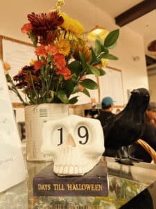 A countdown calendar shaped like a skull shows "19 days till Halloween," accompanied by a colorful bouquet of autumn flowers and a decorative black crow in Salem Massachusetts