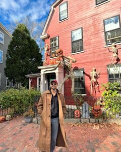 A man in a brown coat and red cap stands in front of a red house decorated with skeletons and pumpkin-headed figures for Halloween in Salem, Massachusetts. The house's yard features tombstones and carved pumpkins.