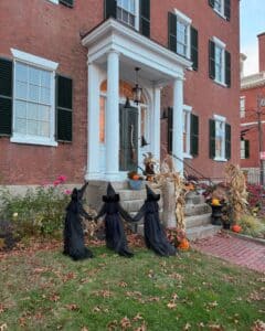 A historic brick home in Salem, Massachusetts, decorated for Halloween with three small witch figures dressed in black, holding hands on the lawn. The porch features pumpkins, corn stalks, and festive autumn decor, with warm light glowing from inside the house.