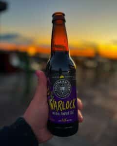 A hand holding a bottle of Southern Tier Warlock Imperial Pumpkin Stout against a backdrop of a sunset sky, with orange and purple hues reflecting off the bottle.