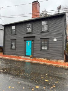 A historic gray wooden house with a bright blue front door and a large brick chimney. The house has a simple design with black window frames and is set against a brick sidewalk on a rainy day in Salem, Massachusetts.