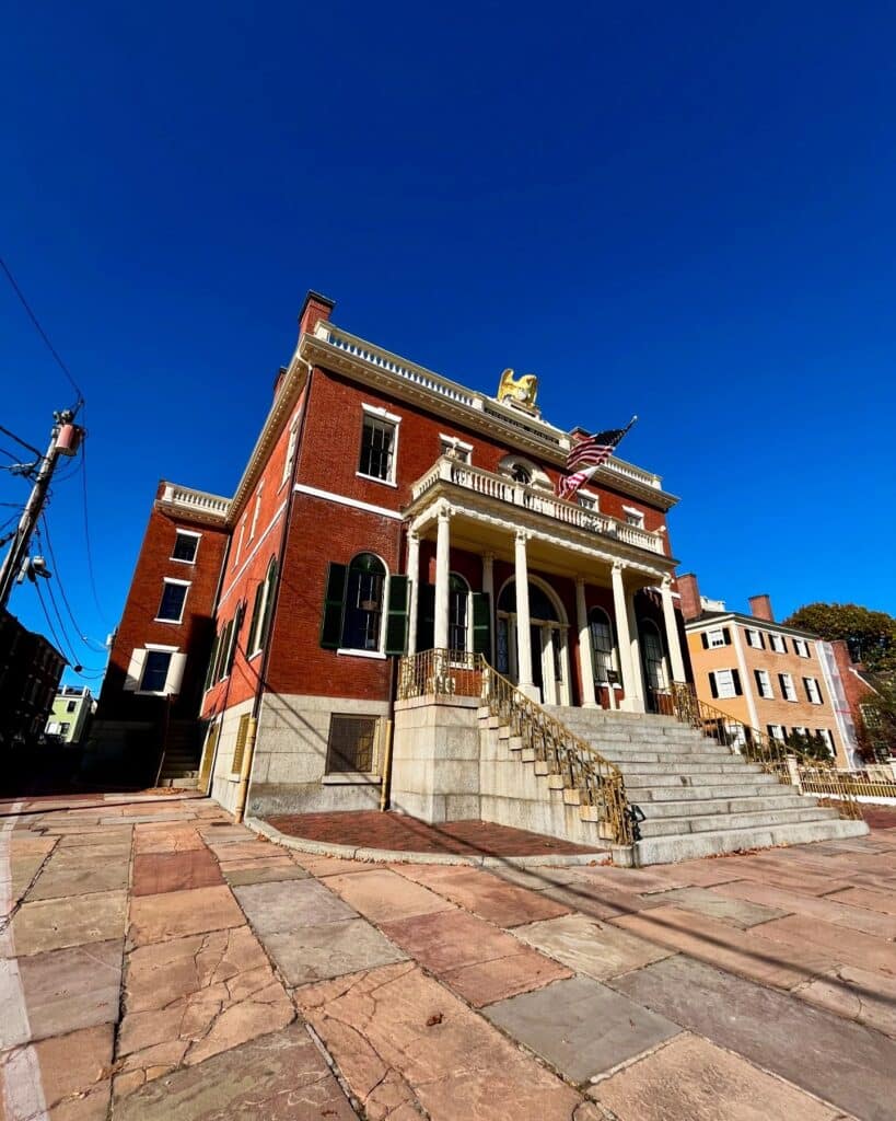 A historic brick building in Salem, Massachusetts, featuring a grand staircase with gold railings, white columns, and arched windows, set against a bright blue sky with a U.S. flag waving above. The stone pavement in the foreground shows weathered charm.