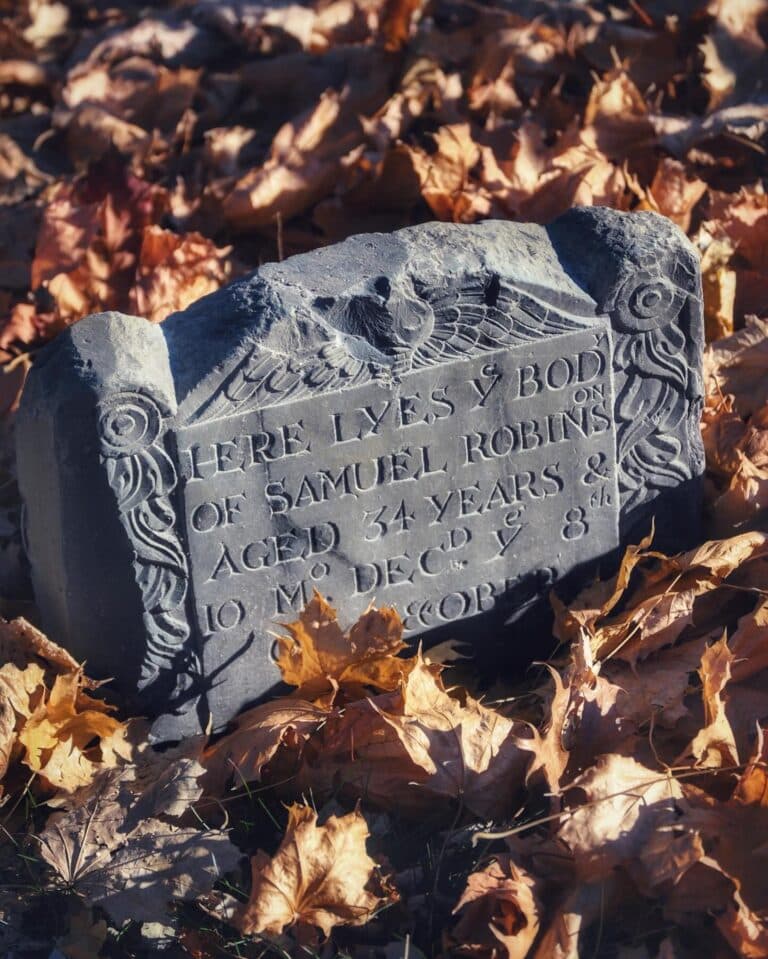 A weathered gravestone of Samuel Robins, partially covered by fallen autumn leaves, stands at Old Burying Point Cemetery in Salem, Massachusetts. The stone features intricate carvings and old-fashioned lettering.