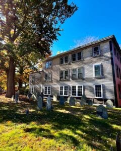 Gravestones in the Old Burying Point Cemetery in Salem Massachusetts, set against the backdrop of a weathered colonial-era building and vibrant autumn trees.
