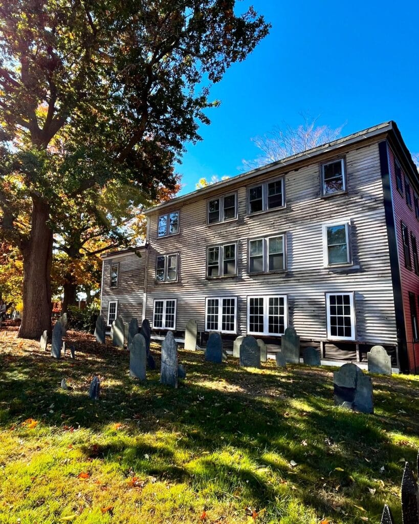 Gravestones in the Old Burying Point Cemetery in Salem Massachusetts, set against the backdrop of a weathered colonial-era building and vibrant autumn trees.