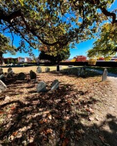 Historic gravestones beneath a large oak tree at the Old Burying Point cemetery in Salem Massachusetts, with dappled sunlight filtering through the branches and autumn leaves scattered on the ground.