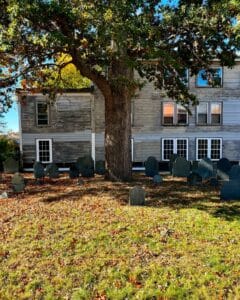 Gravestones and a large oak tree in front of a weathered building at the Old Burying Point Cemetery in Salem, Massachusetts, surrounded by fallen autumn leaves.