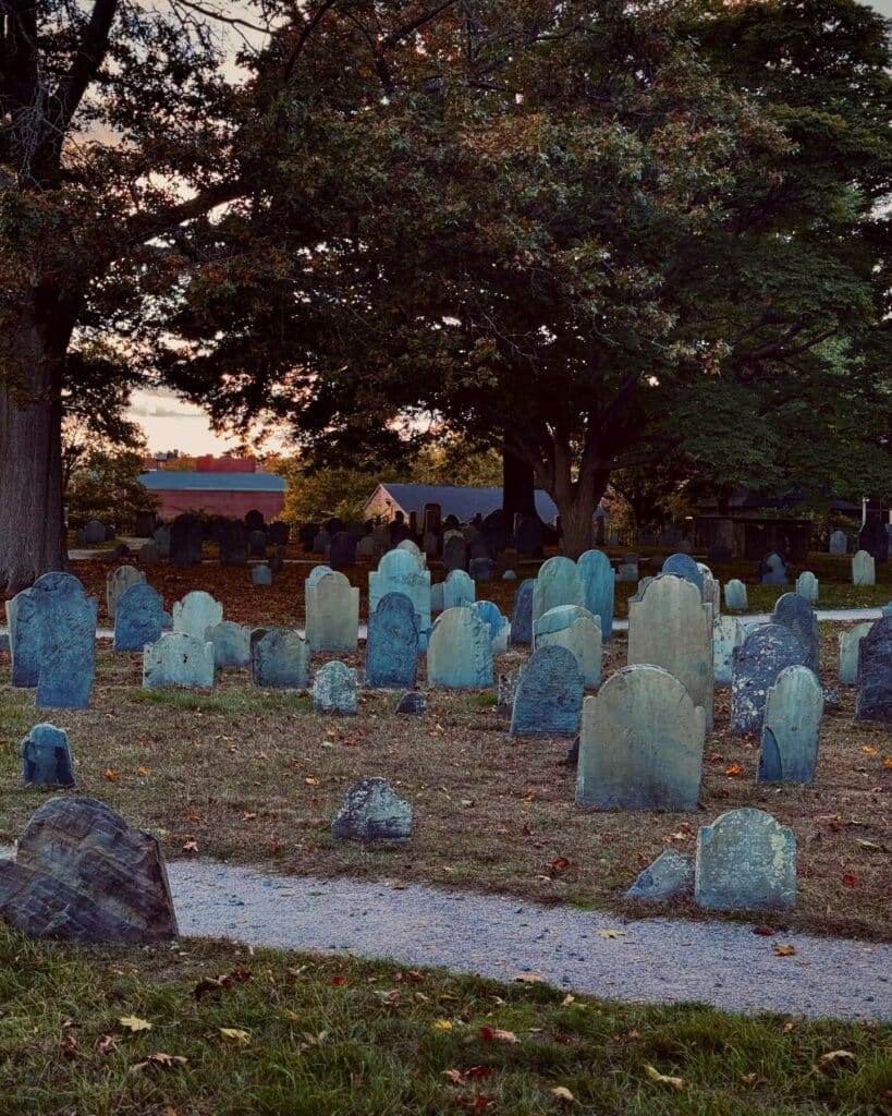 Rows of historic gravestones in the Old Burying Point Cemetery in Salem, Massachusetts, under the shade of large trees with a sunset in the background.