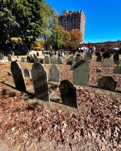 Gravestones at the Old Burying Point Cemetery in Salem, Massachusetts, surrounded by autumn leaves, with trees and a brick building in the background.