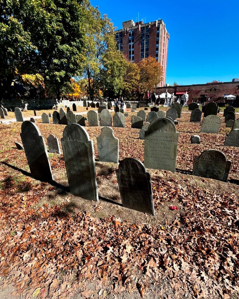 Gravestones at the Old Burying Point Cemetery in Salem, Massachusetts, surrounded by autumn leaves, with trees and a brick building in the background.