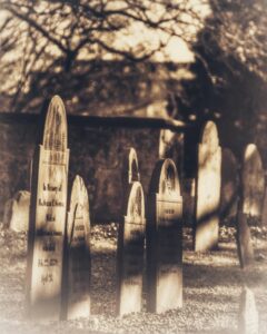 A row of weathered headstones at Old Burying Point Cemetery in Salem, Massachusetts, with soft sunlight and shadows cast by tree branches. The aged stones stand tall in the quiet, historic graveyard.