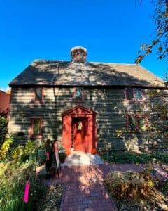 A historic house at the Salem Witch Trials Memorial in Salem, Massachusetts, featuring diamond-paned windows, a red door, and a central brick chimney. The entrance is adorned with pumpkins, hay bales, and seasonal plants under a clear blue sky.