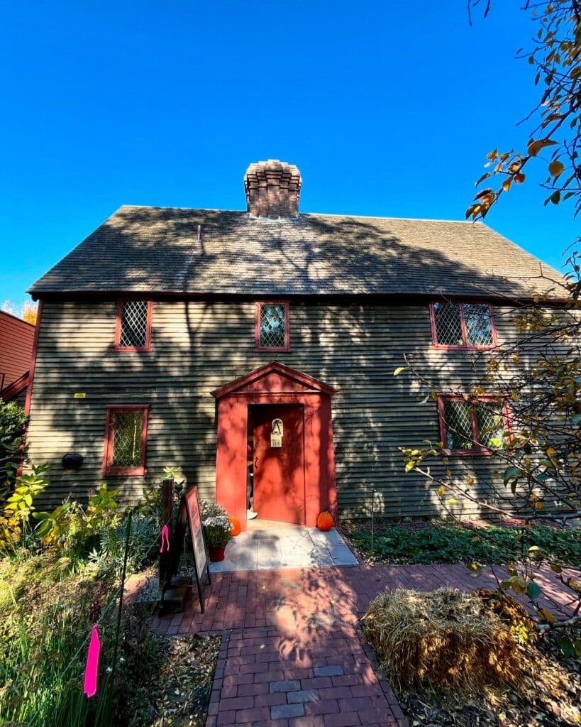  A historic house at the Salem Witch Trials Memorial in Salem, Massachusetts, featuring diamond-paned windows, a red door, and a central brick chimney. The entrance is adorned with pumpkins, hay bales, and seasonal plants under a clear blue sky.