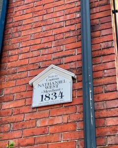 A historic plaque on the red brick wall of the Salem Inn West House in Salem Massachusetts, stating it was built in 1834 for Captain Nathaniel West, a merchant.