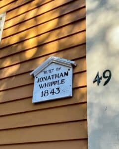 Close-up view of the Jonathan Whipple House marker in Salem, Massachusetts, showing "Built by Jonathan Whipple 1843" on a yellow wood-paneled exterior with the house number 49 displayed nearby.