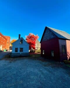 The Quaker Meeting House in Salem, Massachusetts, surrounded by autumn trees, including a vibrant red-leafed tree, under a bright blue sky.