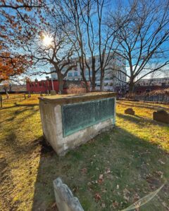 A large stone tomb with a metal plaque stands at Old Burying Point Cemetery in Salem, Massachusetts. Sunlight shines through the trees, casting long shadows across the grass and gravestones.
