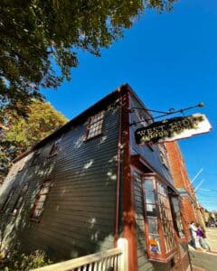 The West India Goods Store in Salem, Massachusetts, featuring its dark green wooden exterior, red-trimmed windows, and a gold hanging sign under a bright blue sky with tree branches framing the scene.