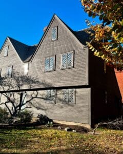Close view of the John Ward House in Salem, Massachusetts, highlighting its dark wood exterior, lattice-style windows, and surrounding autumn foliage under a bright blue sky.