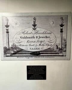 A vintage advertisement for Robert Brookhouse, a goldsmith and jeweler on Essex Street in Salem, Massachusetts, paired with a plaque explaining the evolution of funeral rituals in the 1800s.