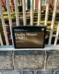 Historical marker for the Derby House in Salem, Massachusetts, showing its establishment in 1762 as part of the Salem Maritime National Historic Site, with the house visible in the background through a white fence.