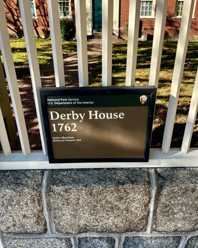 Historical marker for the Derby House in Salem, Massachusetts, showing its establishment in 1762 as part of the Salem Maritime National Historic Site, with the house visible in the background through a white fence.