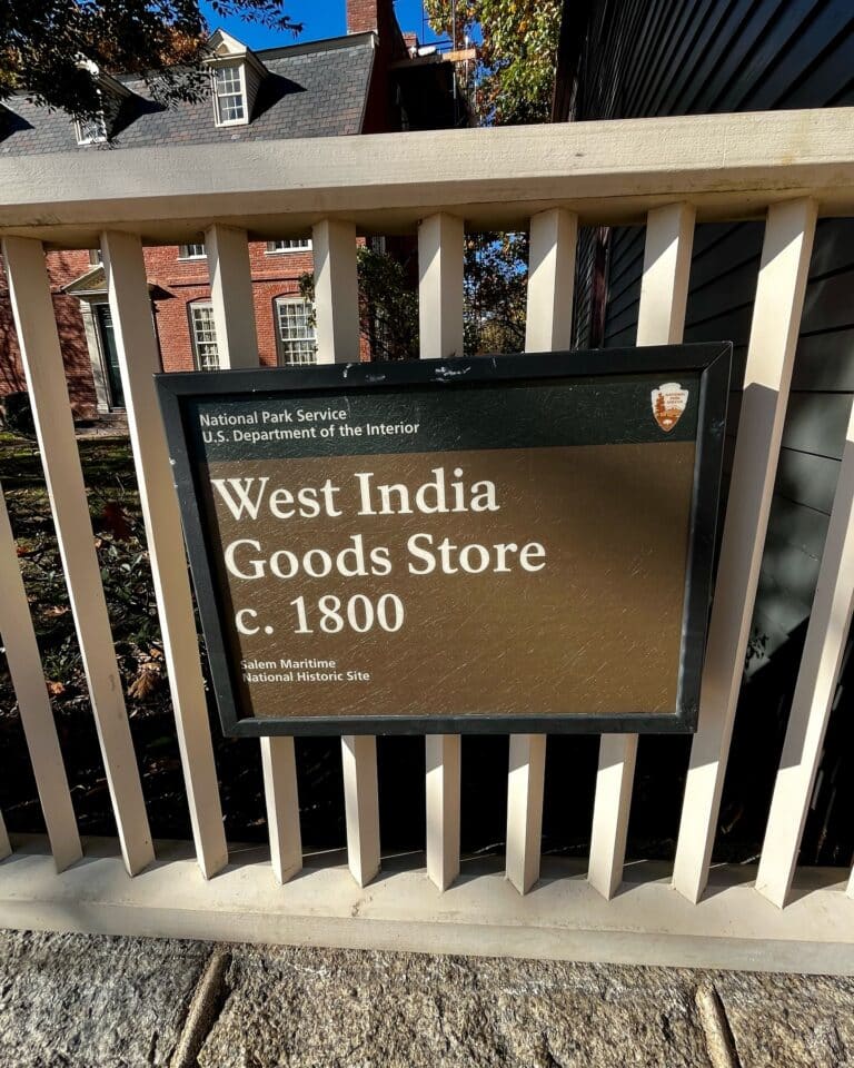 Historical marker for the West India Goods Store in Salem, Massachusetts, dating to circa 1800, located within the Salem Maritime National Historic Site, with a white picket fence and historic buildings visible in the background.