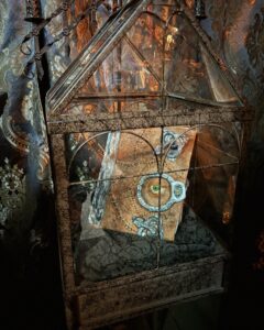 A replica of the Hocus Pocus spellbook, featuring a lifelike eye and stitched leather cover, sits inside a glass display case. The case is set against ornate, patterned fabric with warm lighting.
