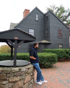 The House of the Seven Gables in Salem Massachusetts featuring a black colonial-style home with a stone well in the foreground and a person standing nearby.