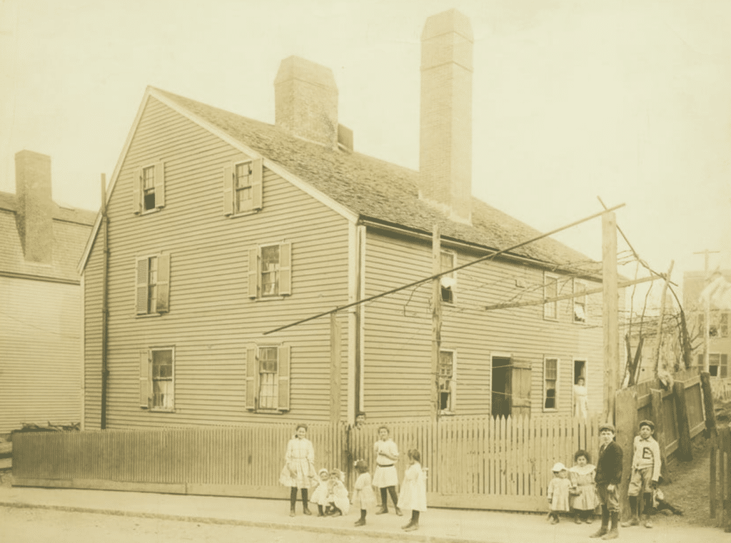 This historic photograph shows the Gedney House in Salem, Massachusetts, as it appeared in the late 19th or early 20th century. The clapboard exterior, tall chimneys, and steeply pitched roof are visible, reflecting the house’s colonial origins. Surrounding the house is a picket fence, and in the foreground, a group of children and young adults pose for the photograph, giving a glimpse into the community life of the time. The image captures a moment when the house was likely being used as a boarding house, highlighting its evolution from a 17th-century home to a multi-purpose residence in a growing urban setting.