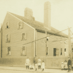 This historic photograph shows the Gedney House in Salem, Massachusetts, as it appeared in the late 19th or early 20th century. The clapboard exterior, tall chimneys, and steeply pitched roof are visible, reflecting the house’s colonial origins. Surrounding the house is a picket fence, and in the foreground, a group of children and young adults pose for the photograph, giving a glimpse into the community life of the time. The image captures a moment when the house was likely being used as a boarding house, highlighting its evolution from a 17th-century home to a multi-purpose residence in a growing urban setting.