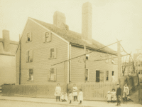 This historic photograph shows the Gedney House in Salem, Massachusetts, as it appeared in the late 19th or early 20th century. The clapboard exterior, tall chimneys, and steeply pitched roof are visible, reflecting the house’s colonial origins. Surrounding the house is a picket fence, and in the foreground, a group of children and young adults pose for the photograph, giving a glimpse into the community life of the time. The image captures a moment when the house was likely being used as a boarding house, highlighting its evolution from a 17th-century home to a multi-purpose residence in a growing urban setting.