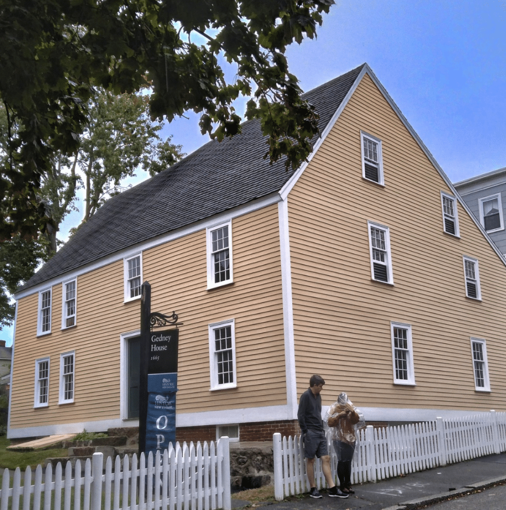 This photo features the Gedney House, a 1665 First Period colonial home located in Salem, Massachusetts. The yellow clapboard siding, steeply pitched roof, and white-trimmed windows are hallmarks of its historic design. A sign in front identifies it as part of Historic New England, inviting visitors to explore its preserved interior and learn about colonial life and architecture. A white picket fence frames the property, adding to its charm. Two visitors are seen in the foreground, emphasizing its status as a popular educational destination for those interested in Salem’s rich history beyond the witch trials.