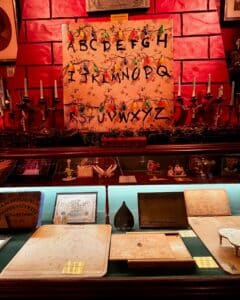 A vibrant display at the Salem Witch Board Museum in Salem, Massachusetts, featuring Ouija boards, a "Stranger Things"-inspired alphabet wall with string lights, and various artifacts in a glass case illuminated with warm lighting.