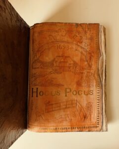 Interior page of the "Hocus Pocus" spellbook, showing artwork with the year 1693, a house illustration, and decorative symbols.