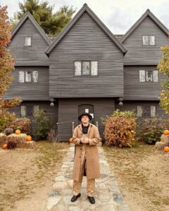 A person wearing a long tan coat and wide-brimmed hat stands confidently on a stone pathway leading to the Salem Witch House, surrounded by autumnal decor including pumpkins and colorful foliage.