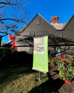 The John Ward House in Salem Massachusetts with a green informational sign in the foreground, surrounded by vibrant red flowers and a clear blue sky.