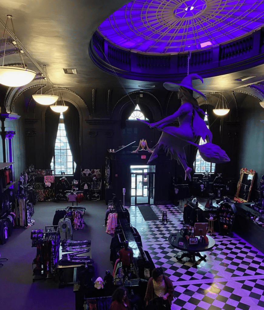 The gothic interior of Blackcraft Salem in Salem, Massachusetts, showcasing a suspended witch sculpture flying on a broom, illuminated by purple lighting, with black walls, checkered floors, and retail displays.
